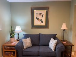 a sage green office with a blue love seat with a large print of a magnolia above it, and tables with lamps and plants on either side.