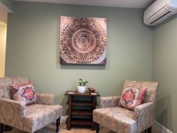 An image of two wing backed chairs in front of a large mandala in a sage green office with a small table and plants.