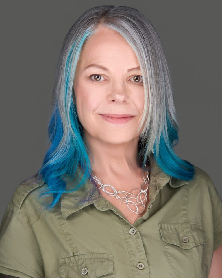 An image of Bobbie Winter, therapist, smiling. She has grey-white hair with the under part died a brilliant blue. She is smiling with a sage green shirt on and a multi-tiered silver necklace 